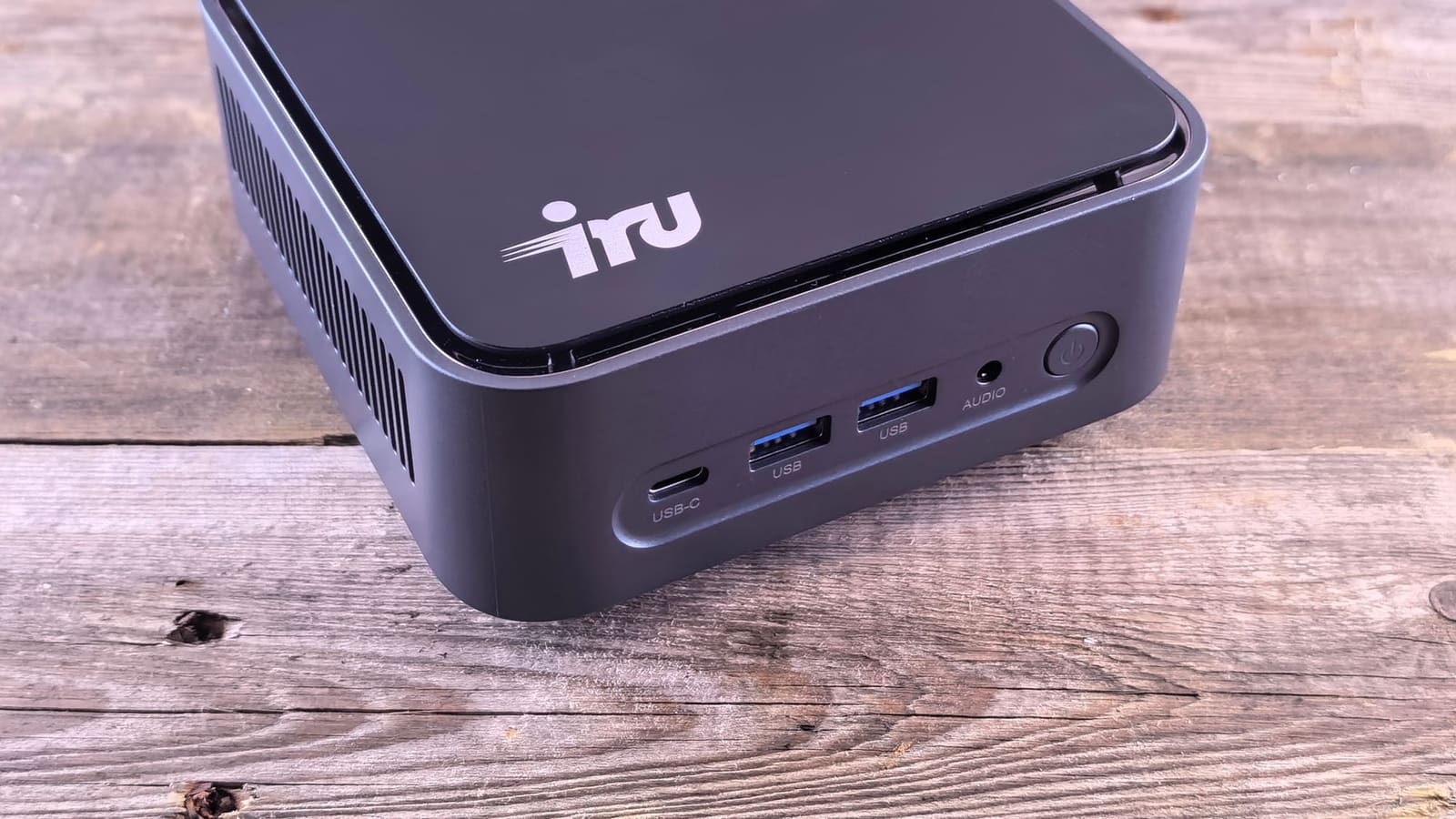 Small black mini PC with USB ports on its front edge, sitting on a weathered wooden desk