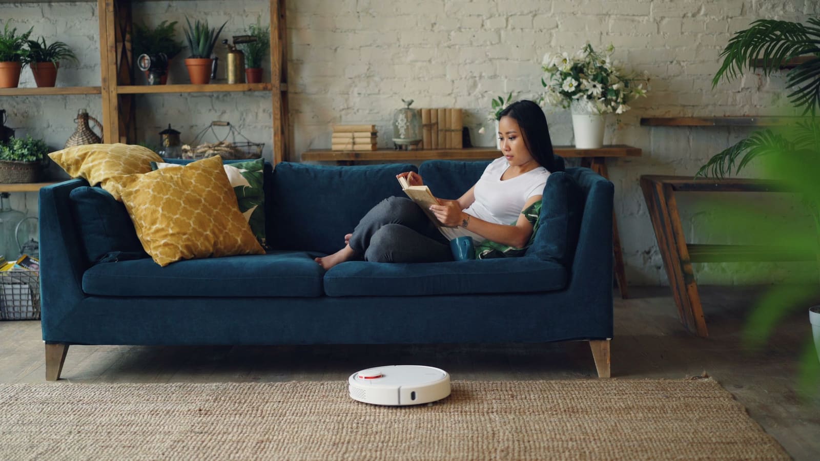 Woman reading on a blue sofa while a white robot vacuum cleans the rug in front of her