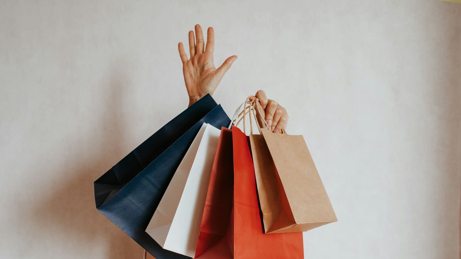 Person holding up several navy, white, red and brown paper shopping bags against a plain wall