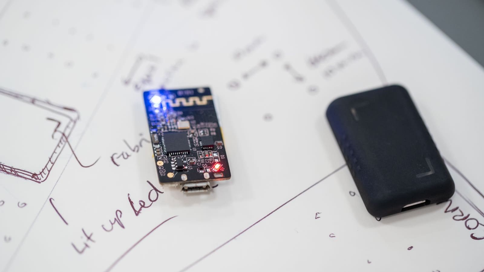 Small black Bluetooth circuit board with an etched antenna and glowing LEDs beside a dark enclosure on a sketched worktop