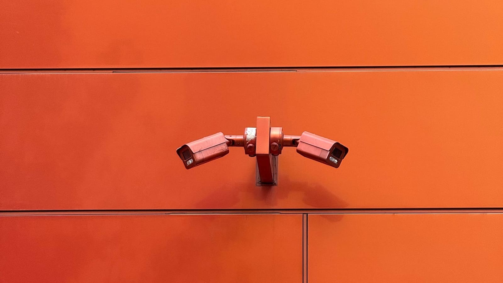 Two white security cameras mounted side by side on a bright orange wall