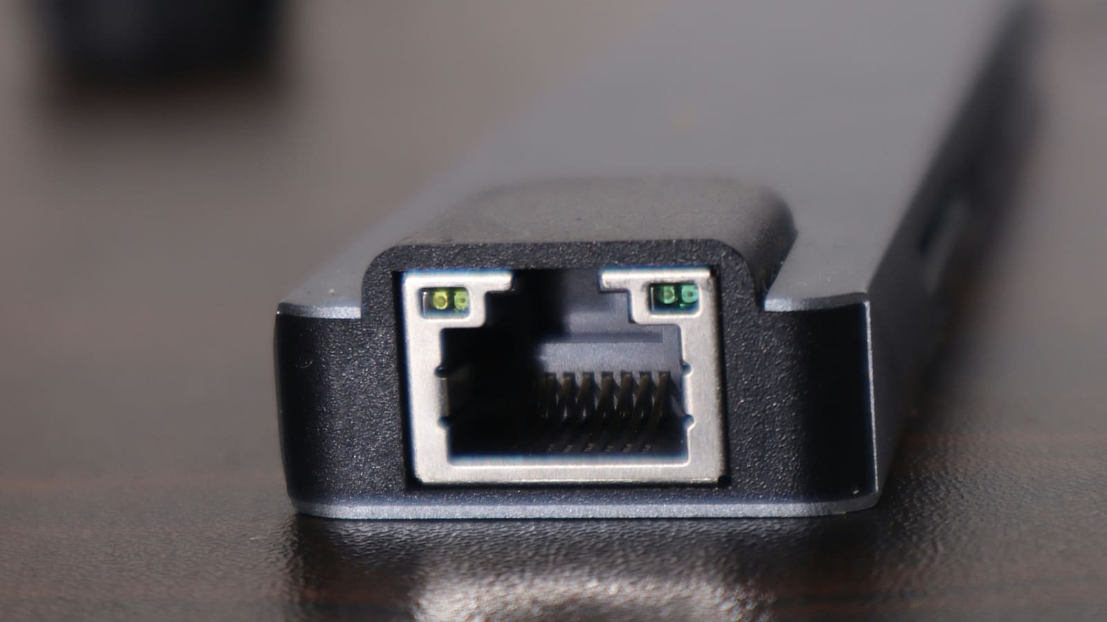 Close-up of an Ethernet port with green link lights on a slim grey network adapter on a dark desk.